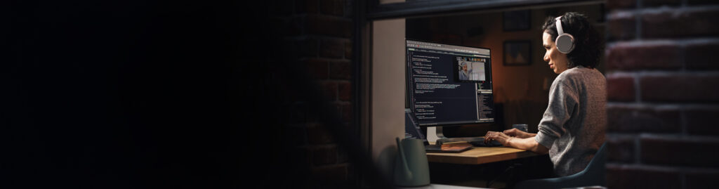 A person wearing headphones working on a computer, displaying code and a video call, in a dimly lit room.