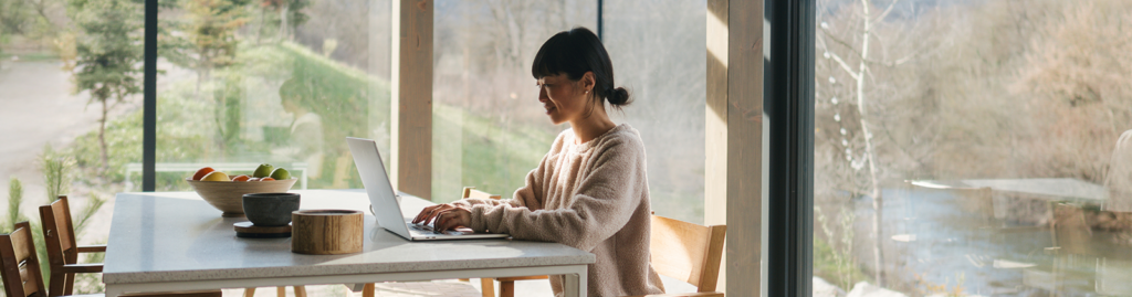 A woman focused on her laptop in a bright, minimalist room, enjoying a peaceful view of trees and nature through floor-to-ceiling windows.